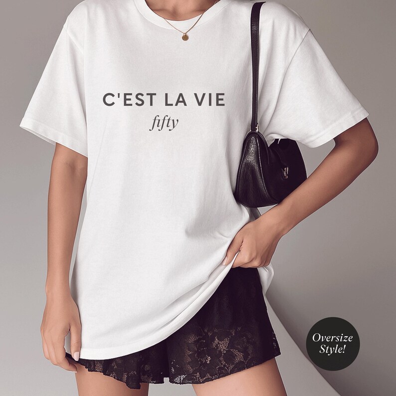 May include: White oversized t-shirt with the text "C'EST LA VIE fifty" in black. The shirt is paired with black lace shorts and a small black shoulder bag. A gold necklace is also visible.