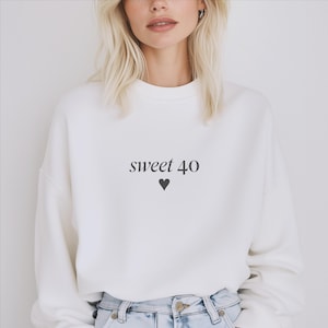 Könnte beinhalten: Ein weißes Sweatshirt mit Rundhalsausschnitt und dem Schriftzug "sweet 40" in einer dunkelgrauen Serifenschrift, platziert über einem kleinen, ausgefüllten dunkelgrauen Herzsymbol. Das Kleidungsstück hat lange Ärmel und eine lockere Passform.