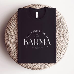 Könnte beinhalten: Schwarzes T-Shirt mit dem weißen Schriftzug "ALL I THINK ABOUT IS KARMA". Das Design umfasst einen halbkreisförmigen Bogen, Sterne und eine Weltkugel. Das T-Shirt wird auf einer gewebten, kreisförmigen Oberfläche präsentiert.