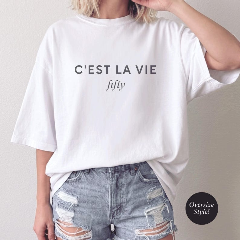 May include: White oversized t-shirt with the text "C'EST LA VIE fifty" in black. The shirt is paired with denim shorts. A black circle with the words "Oversize Style!" is in the lower right corner.