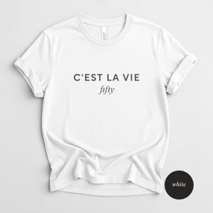 May include: White t-shirt with the text "C'EST LA VIE" above the word "fifty" in a script font. The shirt has short sleeves and a crew neck. The shirt is made of a soft, comfortable material.