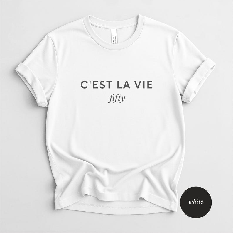 May include: White t-shirt with the text "C'EST LA VIE" above the word "fifty" in a script font. The shirt has short sleeves and a crew neck. The shirt is made of a soft, comfortable material.