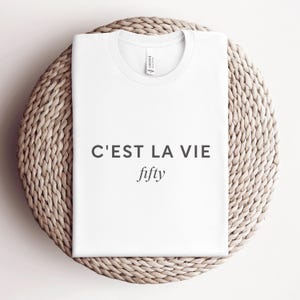 May include: A white t-shirt with the text "C'EST LA VIE" above the word "fifty" in a simple, elegant font. The shirt is neatly folded and placed on a round, woven wicker placemat.