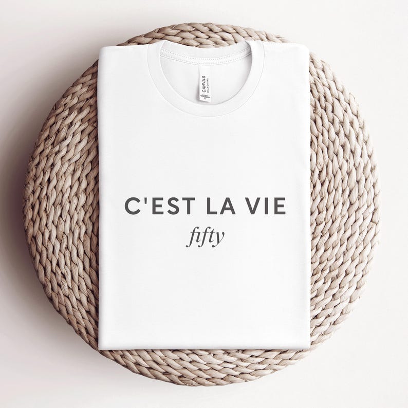 May include: A white t-shirt with the text "C'EST LA VIE" above the word "fifty" in a simple, elegant font. The shirt is neatly folded and placed on a round, woven wicker placemat.