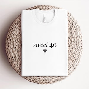Könnte beinhalten: Ein weißes kurzärmeliges T-Shirt mit dem Schriftzug "sweet 40" in einer dunkelgrauen Serifenschrift, platziert über einem kleinen dunkelgrauen Herzsymbol. Das Kleidungsstück hat einen Rundhalsausschnitt und ein sichtbares Markenetikett am Kragen.
