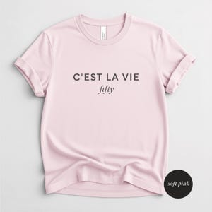 May include: A soft pink t-shirt with rolled-up sleeves. The shirt features the phrase "C'EST LA VIE" above the word "fifty" in a stylish font. The shirt is made of a soft material and is a light pink color.