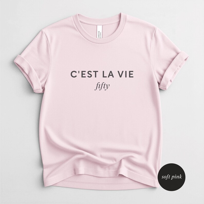 May include: A soft pink t-shirt with rolled-up sleeves. The shirt features the phrase "C'EST LA VIE" above the word "fifty" in a stylish font. The shirt is made of a soft material and is a light pink color.