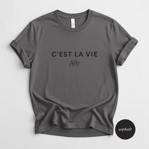 May include: A gray t-shirt with rolled-up sleeves. The text "C'EST LA VIE" is printed above the word "fifty" in a script font. A small black circle with the word "asphalt" is in the lower right corner.