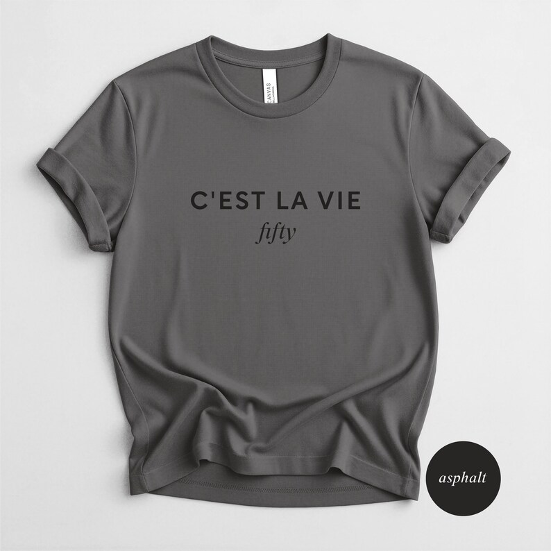 May include: A gray t-shirt with rolled-up sleeves. The text "C'EST LA VIE" is printed above the word "fifty" in a script font. A small black circle with the word "asphalt" is in the lower right corner.