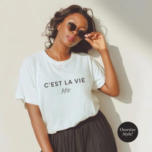 May include: A woman wearing a white t-shirt with the text "C'est La Vie fifty" in black. She is also wearing a black skirt and sunglasses. The image has a soft, natural lighting and a casual, relaxed vibe.