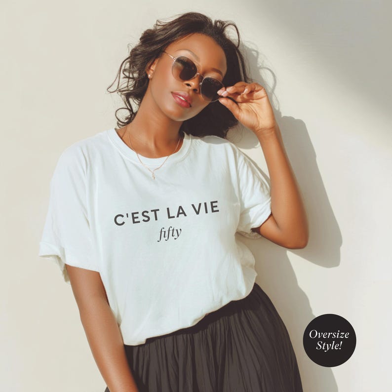 May include: A woman wearing a white t-shirt with the text "C'est La Vie fifty" in black. She is also wearing a black skirt and sunglasses. The image has a soft, natural lighting and a casual, relaxed vibe.
