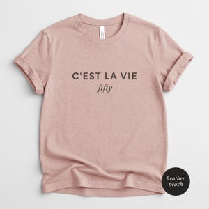 May include: Heather peach-colored t-shirt with rolled sleeves. The shirt features the phrase "C'EST LA VIE" above the word "fifty" in black text. The shirt is made of a soft material and is a casual wear item.