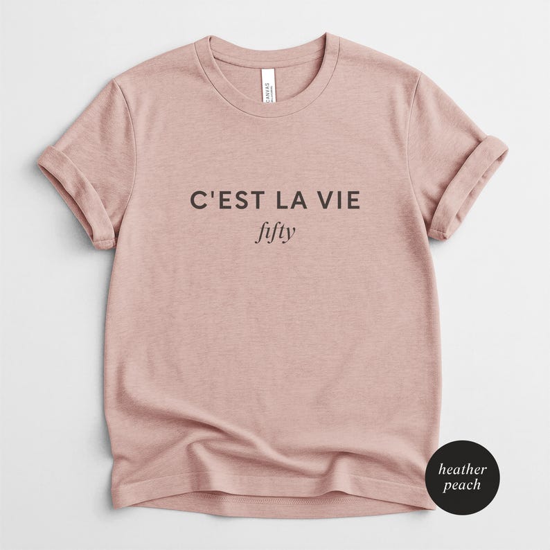 May include: Heather peach-colored t-shirt with rolled sleeves. The shirt features the phrase "C'EST LA VIE" above the word "fifty" in black text. The shirt is made of a soft material and is a casual wear item.