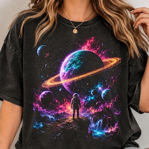 May include: A charcoal black t-shirt featuring a graphic print of an astronaut standing on a rocky terrain looking toward a large planet with orange rings, surrounded by smaller planets, glowing pink and blue nebulae, and distant stars.