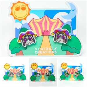 Puede incluir: Un par de pendientes caprichosos con un diseño de personaje de bufón triste, sobre un fondo de circo colorido con un sol y una luna. El texto "CATBAT CREATIONS" es visible.