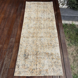 May include: A rectangular runner rug featuring a faded, distressed pattern in shades of tan, beige, and muted blue. The design consists of intricate, repeating floral and geometric motifs spread across the entire surface of the textile.