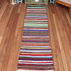 May include: A long, rectangular runner rug featuring a dense, multi-colored horizontal striped pattern. The textile is composed of various woven fabric strips in shades of red, blue, green, yellow, white, and brown, finished with short fringe.