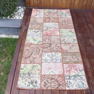 May include: Rectangular patchwork rug with a variety of patterns and colors. The rug features a mix of floral and geometric designs in shades of brown, beige, green, and red. The rug is placed on a wooden deck.
