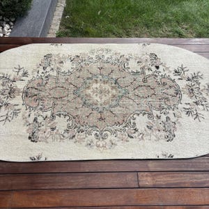Könnte beinhalten: Ein ovaler Teppich mit cremefarbener Basis und einem zentralen Medaillon-Design in gedeckten Tönen von Altrosa, Salbeigrün und Anthrazit. Das komplexe Muster zeigt florale Motive und geschwungene Bordüren auf der gesamten Oberfläche.