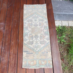 May include: A rectangular runner rug featuring a central diamond medallion pattern in muted shades of sage green, soft orange, and cream. The rug has a symmetrical geometric design with intricate floral-inspired motifs and a fringed edge finish.