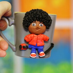 Puede incluir: Una taza de cerámica gris con una figura de arcilla hecha a mano de una persona con piel marrón, cabello negro rizado, vistiendo una chaqueta roja, pantalones azules y zapatos rojos.