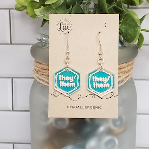 May include: Teal blue hexagonal earrings with the text "they/them" in white lettering. The earrings are hypoallergenic.