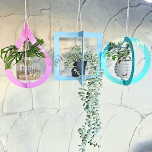 May include: Three hanging planters in various shapes and colours. A pink teardrop-shaped planter holds a clear glass vase with a plant. A blue square planter holds a black pot with greenery. A teal sphere planter holds a white pot with a plant.