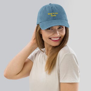 Puede incluir: Gorra de béisbol de mezclilla azul claro con el texto bordado "Sing to me, Paolo" en amarillo. La gorra tiene una visera curva y una correa ajustable. Una persona lleva el sombrero.