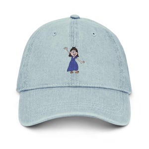 Puede incluir: Gorra de béisbol de mezclilla azul claro con un bordado de dibujos animados de una mujer con un vestido azul saludando. La gorra tiene una visera curva y un botón en la parte superior. El personaje de dibujos animados tiene cabello castaño y usa sandalias.
