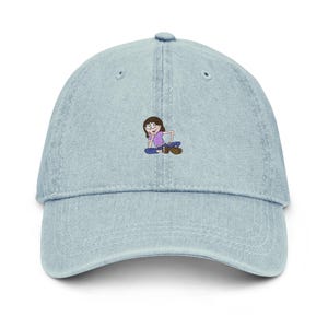 Puede incluir: Gorra de béisbol de mezclilla azul claro con un pequeño gráfico de dibujos animados bordado de una persona sentada. La gorra tiene una visera curva y una correa ajustable en la parte posterior. El gráfico presenta a una persona con gafas, con una camisa morada y pantalones azules.