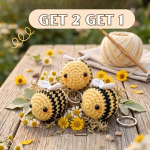 May include: Three handmade crochet bee keyrings with black and yellow stripes, white wings, and black eyes, arranged on a wooden surface with flowers and greenery. The image includes the text "GET 2 GET 1".