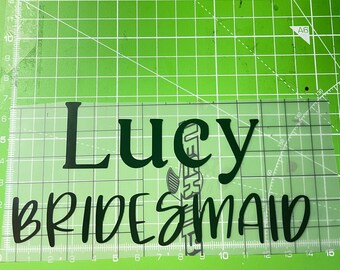 May include: A clear vinyl decal featuring the name "Lucy" and the word "Bridesmaid" in dark green lettering. The decal is positioned on a green cutting mat with grid lines and metric measurements.