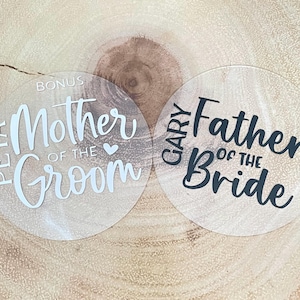 May include: Two clear acrylic wedding coasters. One coaster says "PETA Mother of the Groom" in white script. The other says "GARY Father of the Bride" in black script. Both are round.