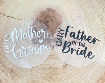 May include: Two clear acrylic wedding coasters. One coaster says "PETA Mother of the Groom" in white script. The other says "GARY Father of the Bride" in black script. Both are round.
