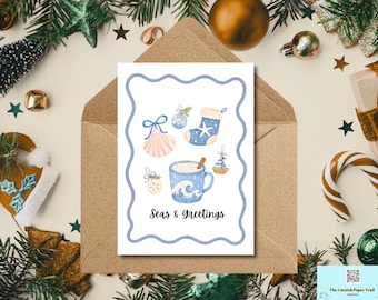 May include: A white greeting card with a wavy blue border featuring illustrations of a seashell, a blue stocking with a starfish, a mug with a wave pattern and cinnamon stick, and a small sailboat. The text reads Seas & Greetings.