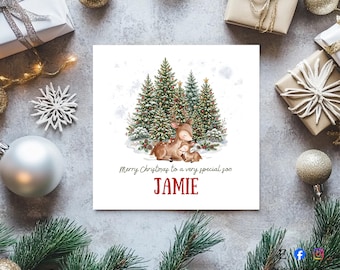 May include: A square greeting card featuring a watercolour illustration of two deer sitting in front of decorated Christmas trees. The text reads Merry Christmas to a very special son and the name JAMIE in red capital letters below the deer.