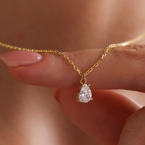 May include: A delicate gold necklace featuring a small, pear-shaped, clear gemstone pendant. The necklace is held by a person's fingers, showcasing the fine chain and the sparkling pendant. The jewelry is set against a blurred background.