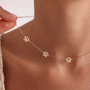 May include: A gold-toned chain necklace featuring three evenly spaced, small paw print charms. Each individual paw print charm is engraved with a name, including the names Cooper, Bubba, and Lola, suspended on a delicate, thin metal link chain.