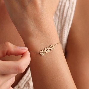May include: Gold-toned bracelet featuring three paw print charms, each with a name engraved: Oreo, Semmy, and Luna. The delicate chain bracelet is being worn on a wrist.