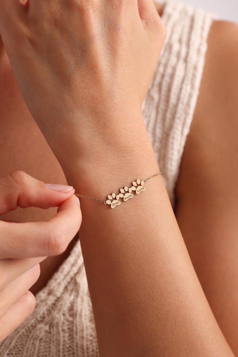 May include: Gold-toned bracelet featuring three paw print charms, each with a name engraved: Oreo, Semmy, and Luna. The delicate chain bracelet is being worn on a wrist.