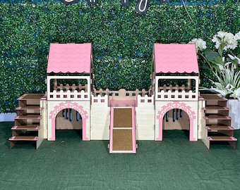 Op de afbeelding: Een houten speelhuis voor kleine huisdieren, met twee roze torens met gebogen ingangen, een helling en twee sets trappen. Het speelhuis is gemaakt van natuurlijk hout en heeft een lichtbruine afwerking. De tekst "Better Together" wordt boven het speelhuis weergegeven.