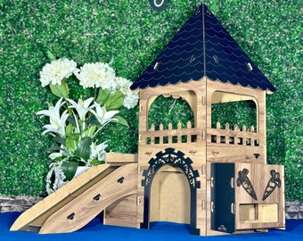Puede incluir: Una casita de juegos de madera para animales pequeños, con un techo negro, un tobogán y una entrada de túnel. La casita tiene una pequeña puerta con un diseño de zanahoria y una valla alrededor de la parte superior.
