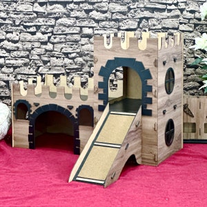 Puede incluir: Una casa de juegos de madera en forma de castillo para animales pequeños, con una rampa, dos entradas y una torre con una ventana. El castillo está hecho de madera marrón con detalles negros.