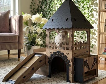 Puede incluir: Un castillo de madera para conejos con techo negro y rampa. El castillo tiene un pequeño conejo blanco en su interior. Cuenta con un arco decorativo y una caja de almacenamiento con zanahorias. El castillo está sobre una alfombra clara.
