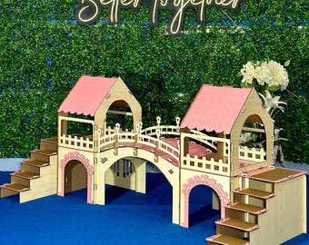 May include: A wooden bridge playset with two pink towers and a curved bridge. The bridge has a white railing and a brown wooden base. The towers have pink roofs and white trim. The playset is on a blue surface. "Better Together" is written in neon letters on a green wall in the background.