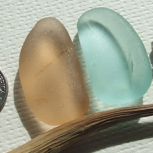 Può includere: Due pezzi di vetro di mare, uno blu chiaro e uno marrone chiaro, con un piccolo ramoscello e una moneta da dieci centesimi di dollaro americano su una superficie bianca testurizzata.
