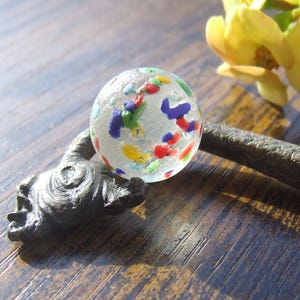 May include: A clear glass marble with colourful flecks of red, blue, yellow, and green rests on a dark, textured, metal sculpture. Yellow flowers are in the background. The marble is the focal point, set against a wooden surface.