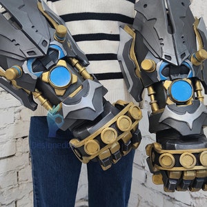 May include: A pair of gray and gold robotic gauntlets with blue accents. The gauntlets have a detailed design with multiple gears and pipes. The gauntlets are worn by a person wearing a white striped sweater and blue jeans.