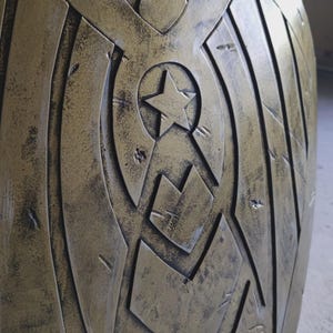 May include: A metallic gold shield with a star and eagle design. The shield has a distressed finish, with a central star emblem and an eagle at the top. The design features geometric shapes and lines.