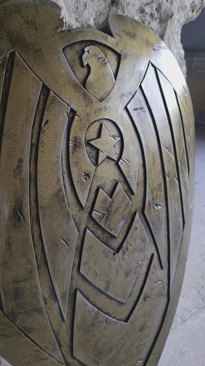 May include: A metallic gold shield with a star and eagle design. The shield has a distressed finish, with a central star emblem and an eagle at the top. The design features geometric shapes and lines.